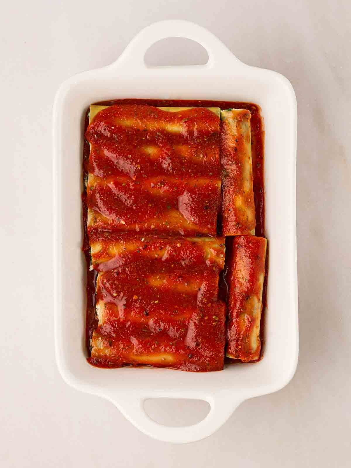 A white oven dish filled with unbaked cannelloni and tomato sauce.