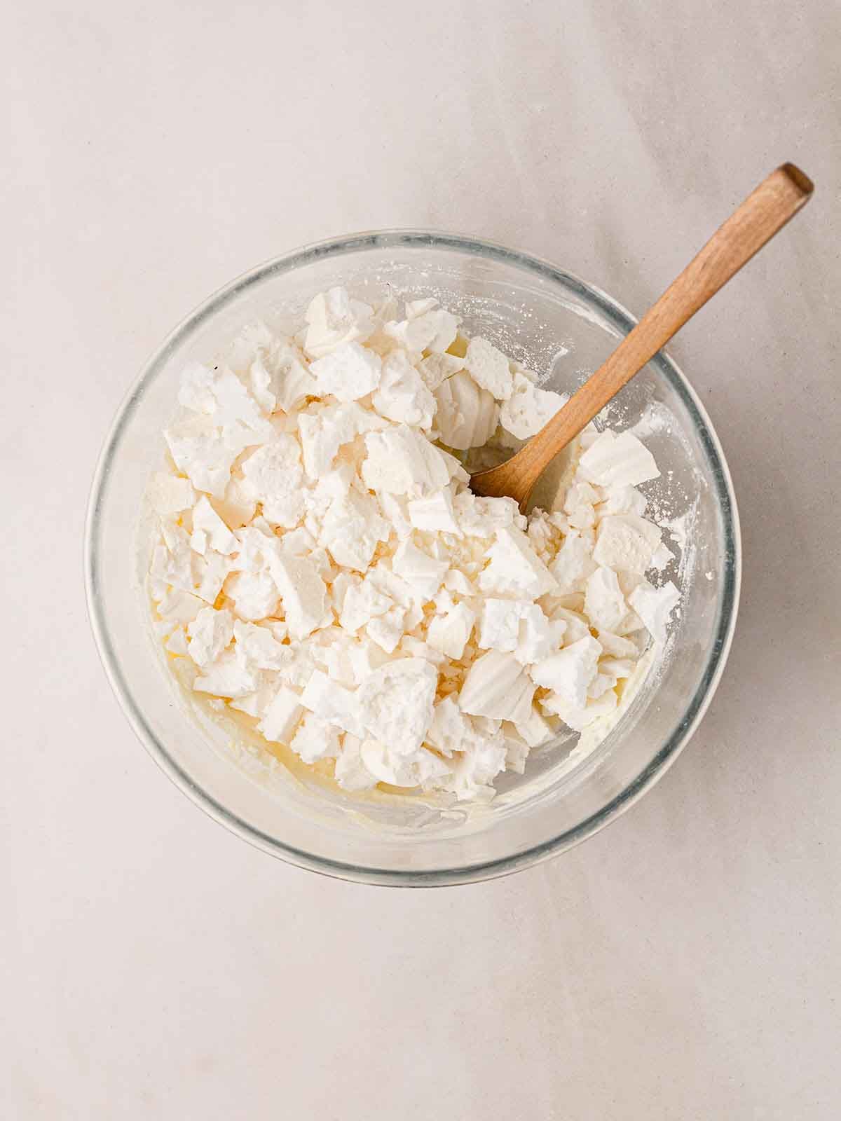 A glass bowl filled with whipped cream and meringue with a wooden spoon sticking out for step 4 in the recipe for Eton Mess.