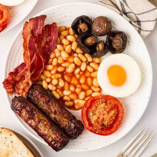 A full English on a white plate, on a white table, with a knife and fork to the side, a plate of toast and a corner of another plate of breakfast.