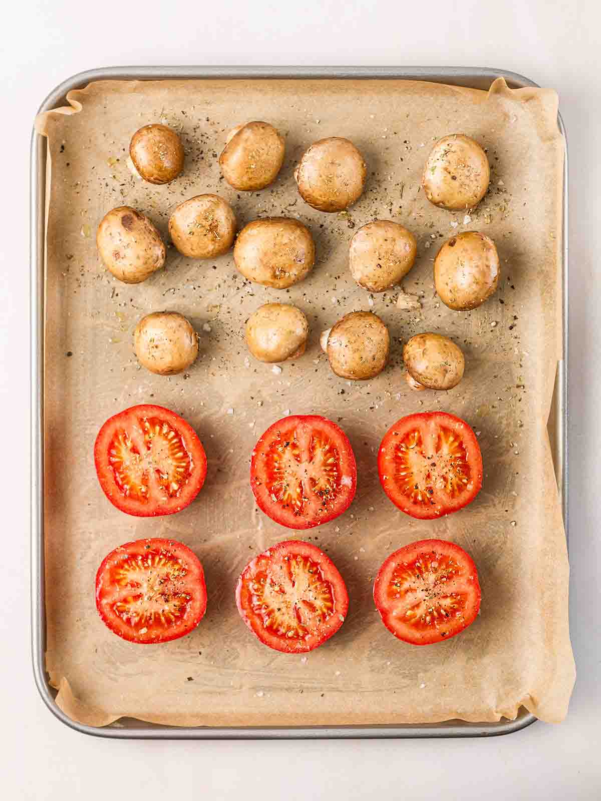 A lined baking tin with halved tomatoes and mushrooms on, seasoned with salt and pepper, for step 2 in the recipe for a full English breakfast.