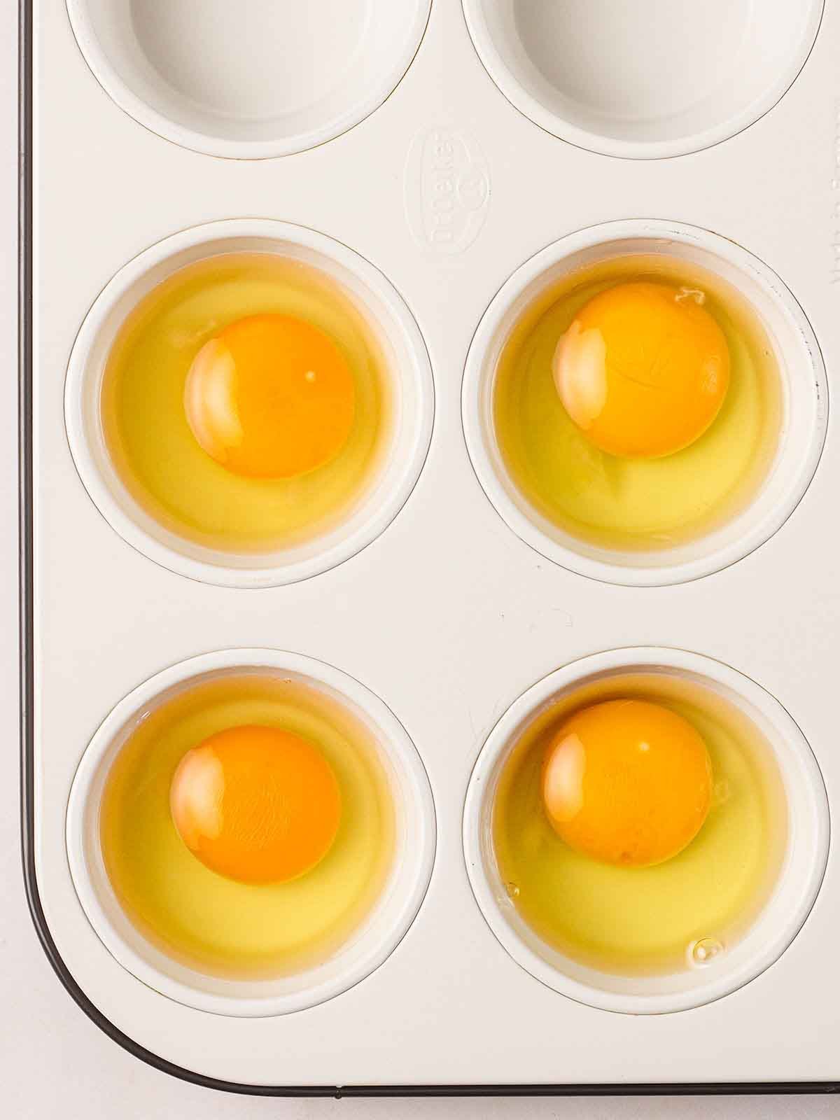 A muffin tray with a raw, cracked egg in each of the 4 holes, for step 4 in the recipe for Full English Breakfast.