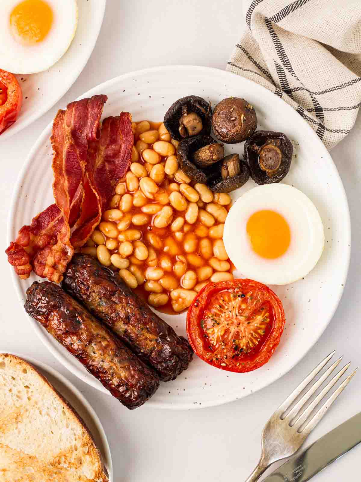 A table with a full English breakfast on, with toast and cutlery to the side.
