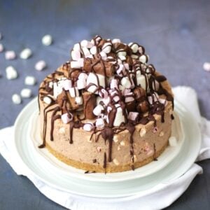 A Rocky Road Cheesecake on a white plate, piled high with marshmallow and chocolate bar topping and melted chocolate.
