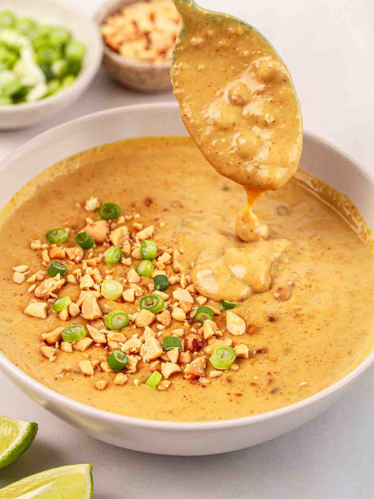 A spoon dipping into a white bowl filled with homemade Satay Sauce.