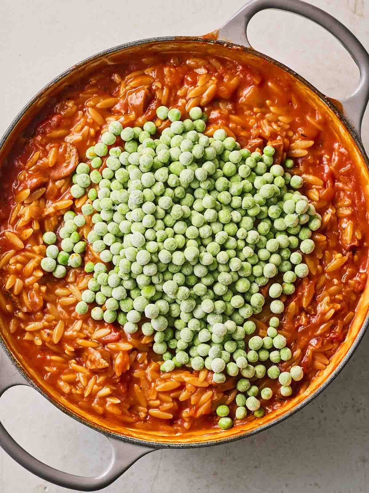 A pan filled with almost-cooked Chorizo Orzo, with frozen peas going in for step 4 in this easy one pot meal.