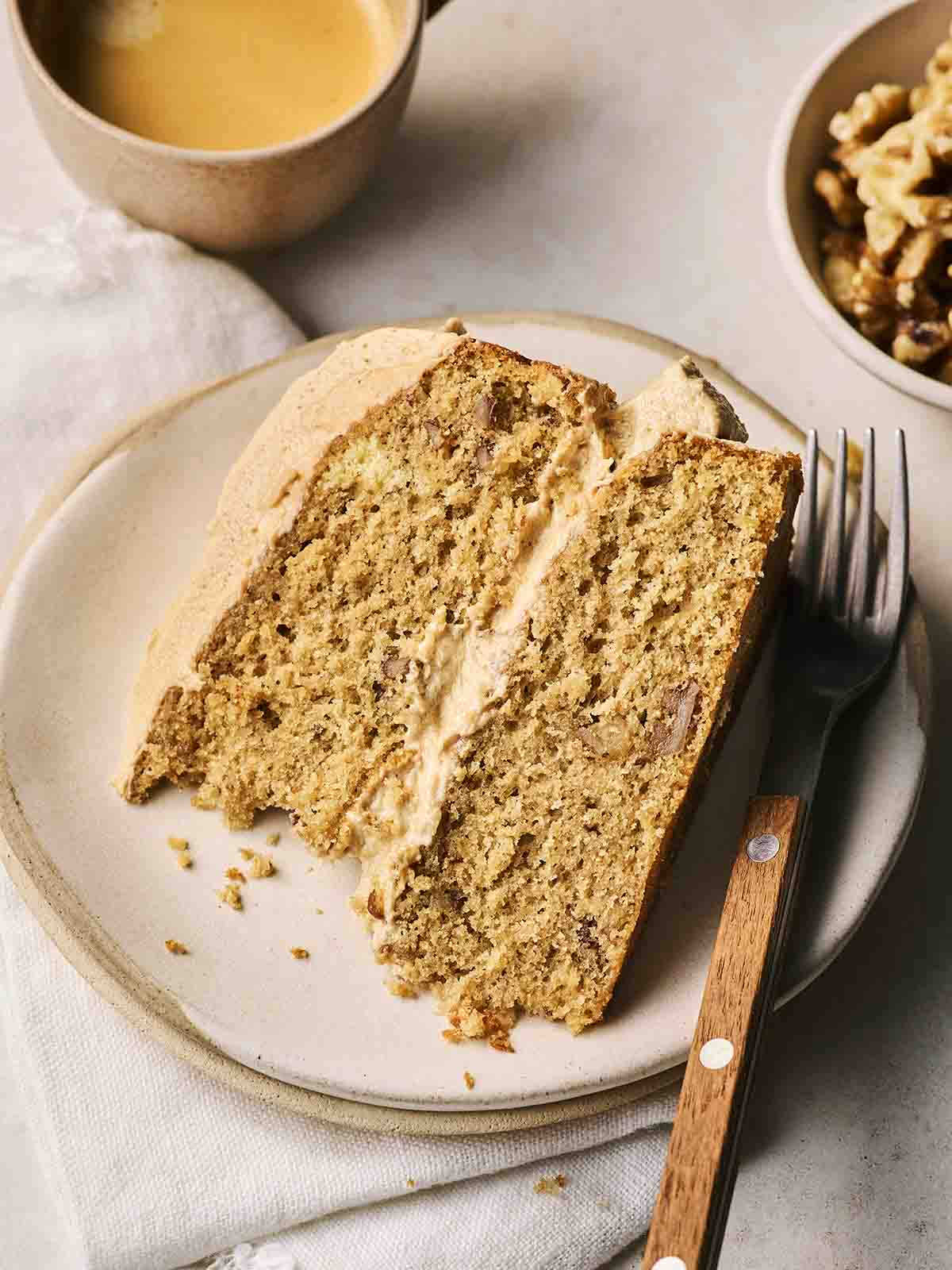 A slice of Coffee and Walnut Cake on a plate, with a fork to the side and a cup of coffee in the background.
