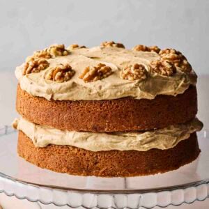A side view of a classic Coffee and Walnut Cake, with two layers of sponge and coffee buttercream and nuts on top, on a glass cake stand.