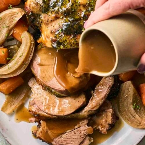 A leg of lamb straight out of the slow cooker, on a plate, with a hand pouring over the gravy, ready to serve.