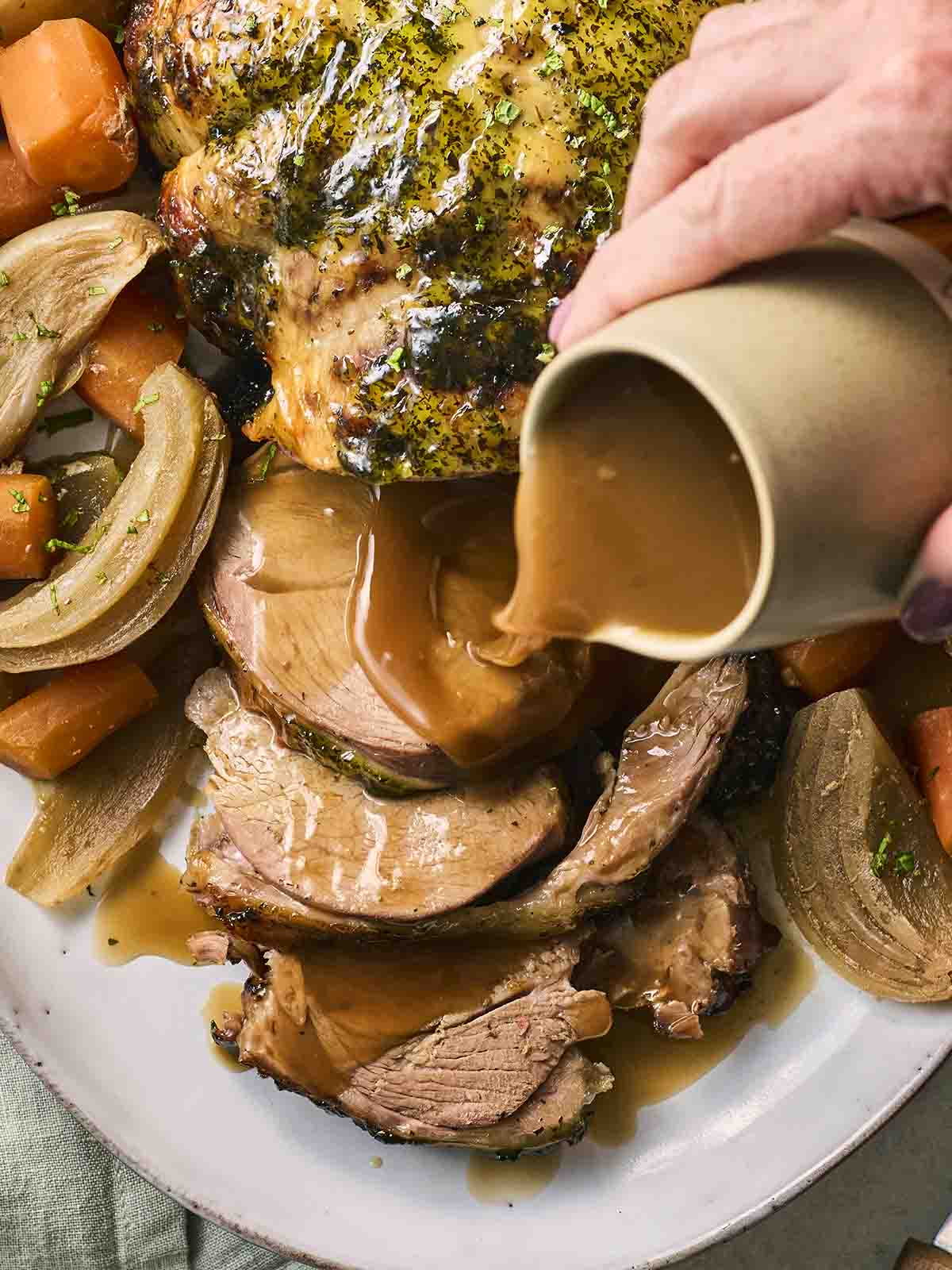 A leg of lamb straight out of the slow cooker, on a plate, with a hand pouring over the gravy, ready to serve.