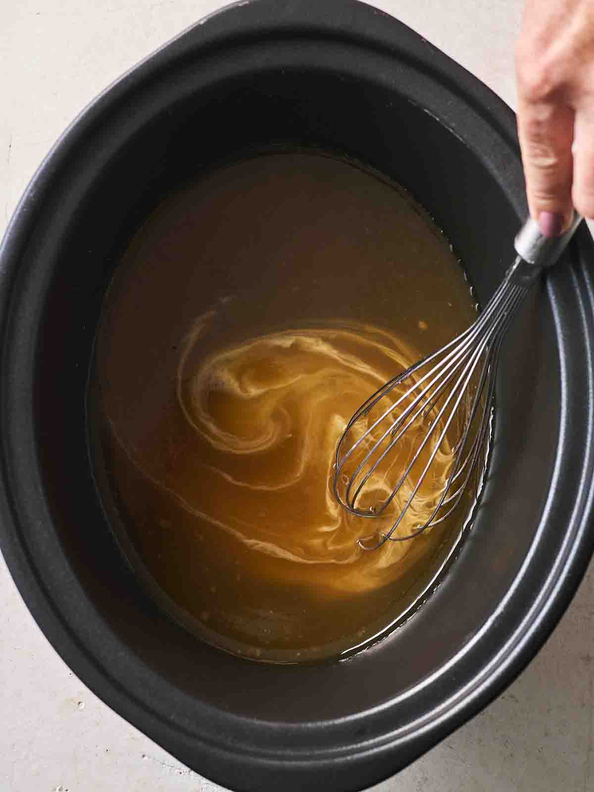 A slow cooker pan with gravy in the bottom, with thickening cornflour stirring in with a whisk, for the recipe Slow Cooker Lamb Leg.