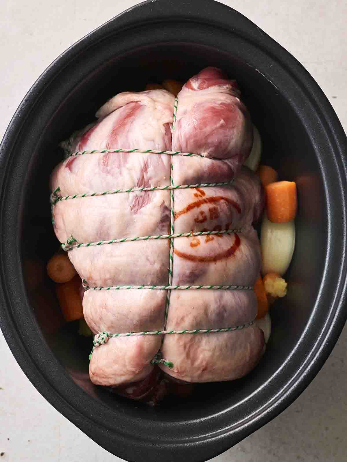A raw leg of lamb in a slow cooker with vegetables underneath.