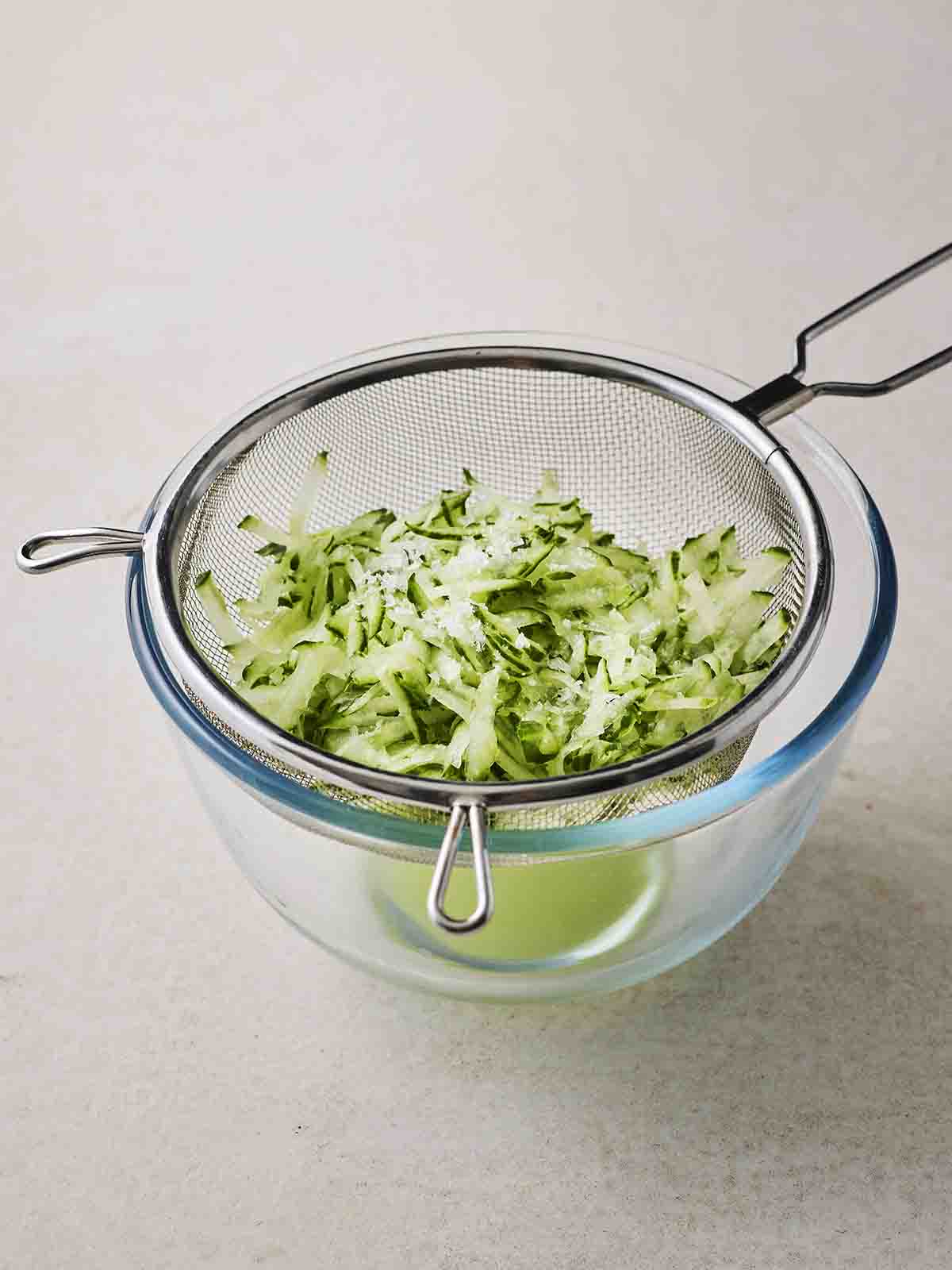 A sieve with grated cucumber inside, over a glass bowl with liquid drained into it, for step 1 in the recipe for Tzatziki.