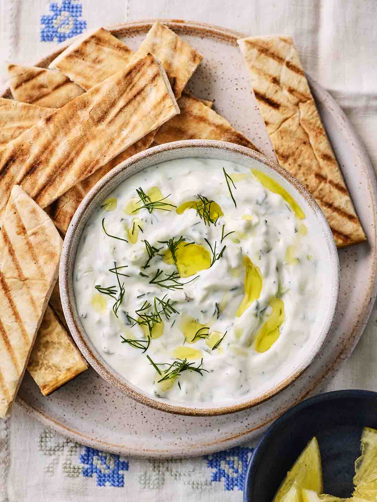 A bowl of Greek Tzatziki dip with olive oil and herbs on top, with sliced pittas to the side.