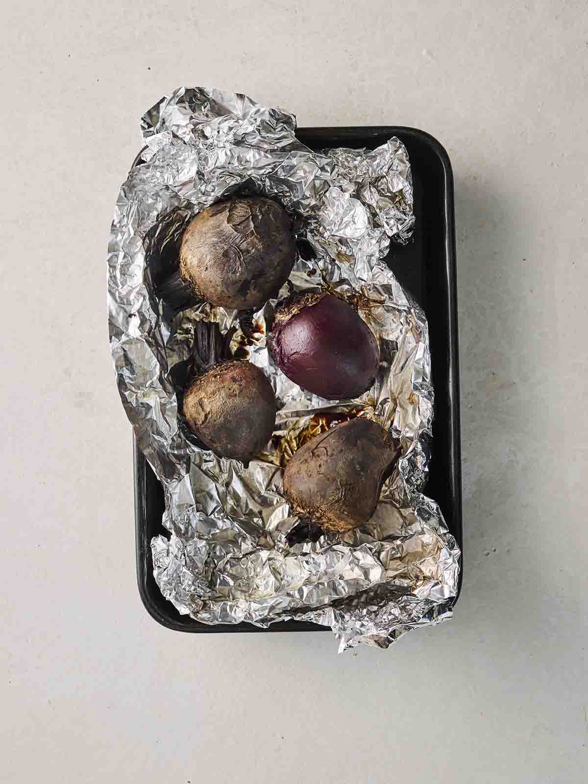 Four whole raw beetroots in top of foil in a baking tin, ready for roasting.