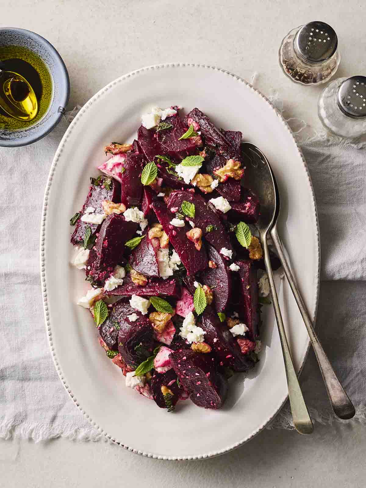 A white serving dish with a homemade Beetroot Salad on, with serving cutlery and dressing to the side.