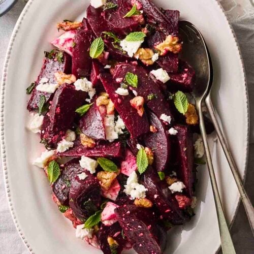 A salad with beetroot, feta, mint and walnuts on a white serving plate with serving spoon and fork.