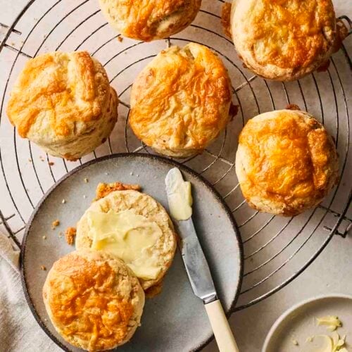 Just-baked Cheese Scones on a wire rack, with one on a plate having been buttered.