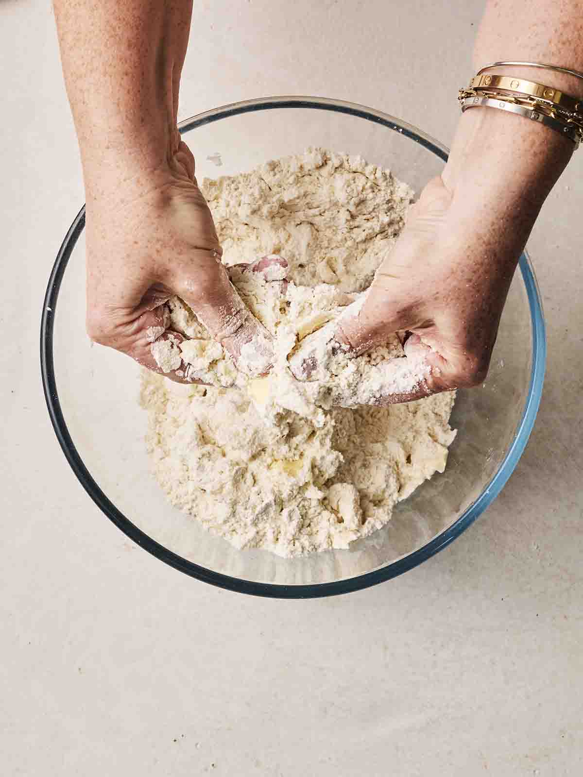 Two hands rubbing flour and butter together over a glass bowl for step 1 in the recipe for Cheese Scones.