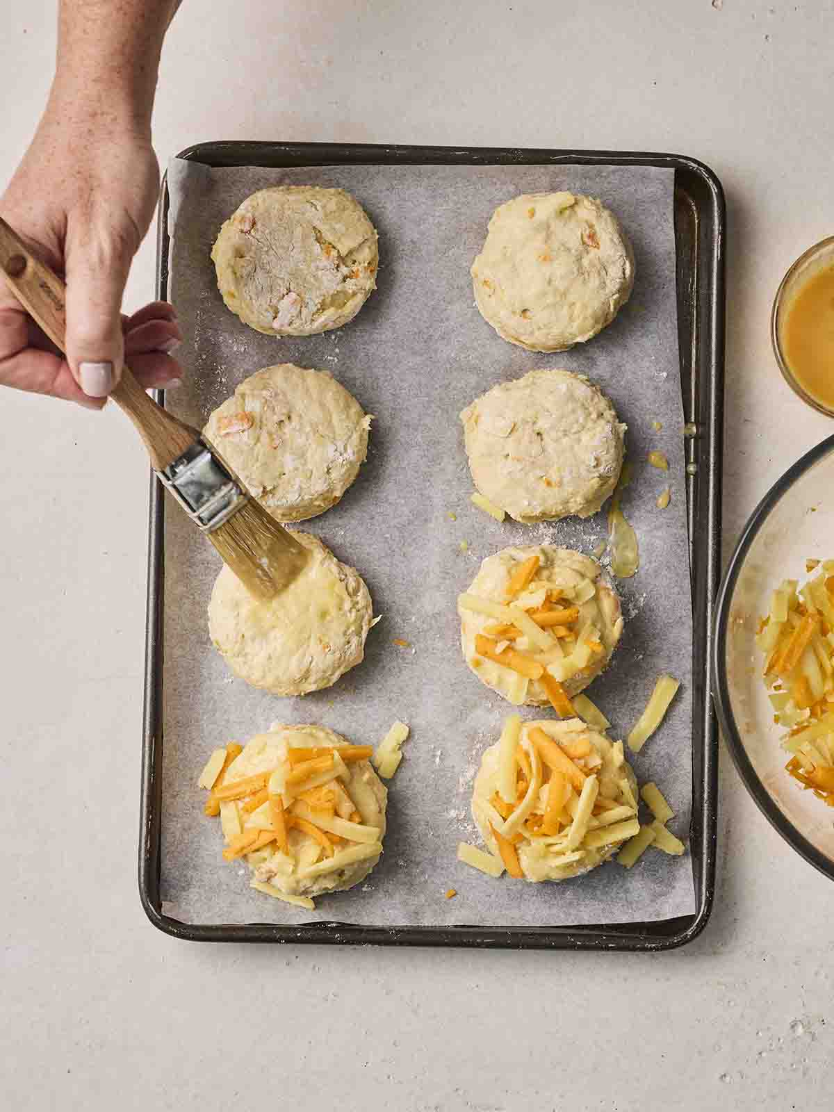 Uncooked Cheese Scones on a baking sheet with a hand brushing on egg.