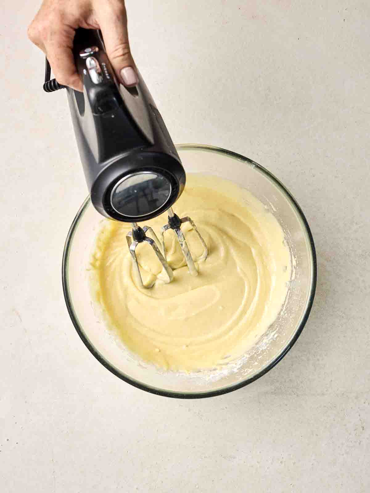 Cake batter in a glass bowl being mixed together with an electric whisk.