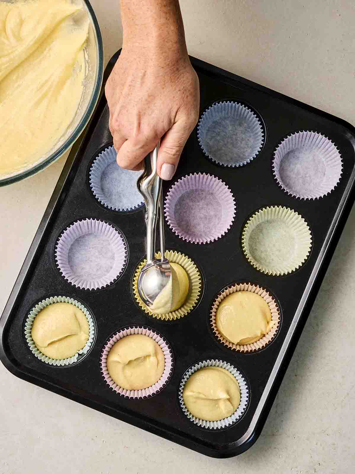 A muffin tray lined with muffin cases, with a hand spooning in cake mixture.
