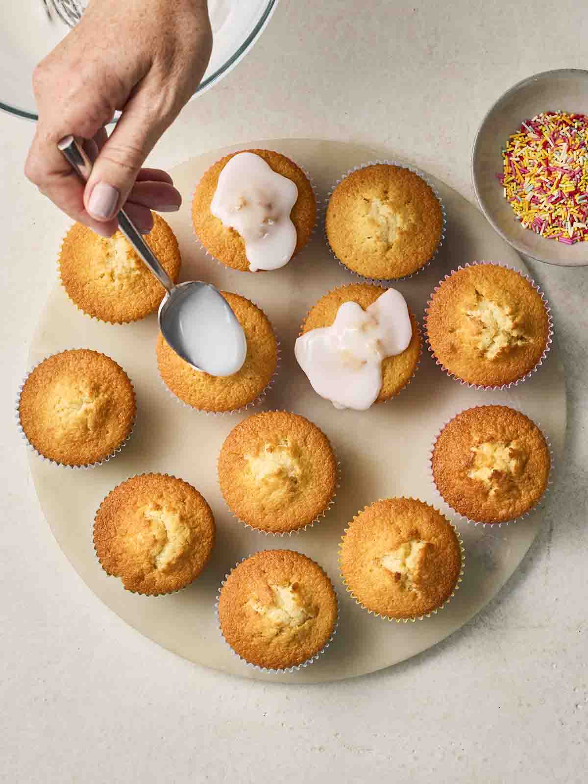 Just-baked Fairy Cakes on a plate with icing being drizzled over the top with a spoon.