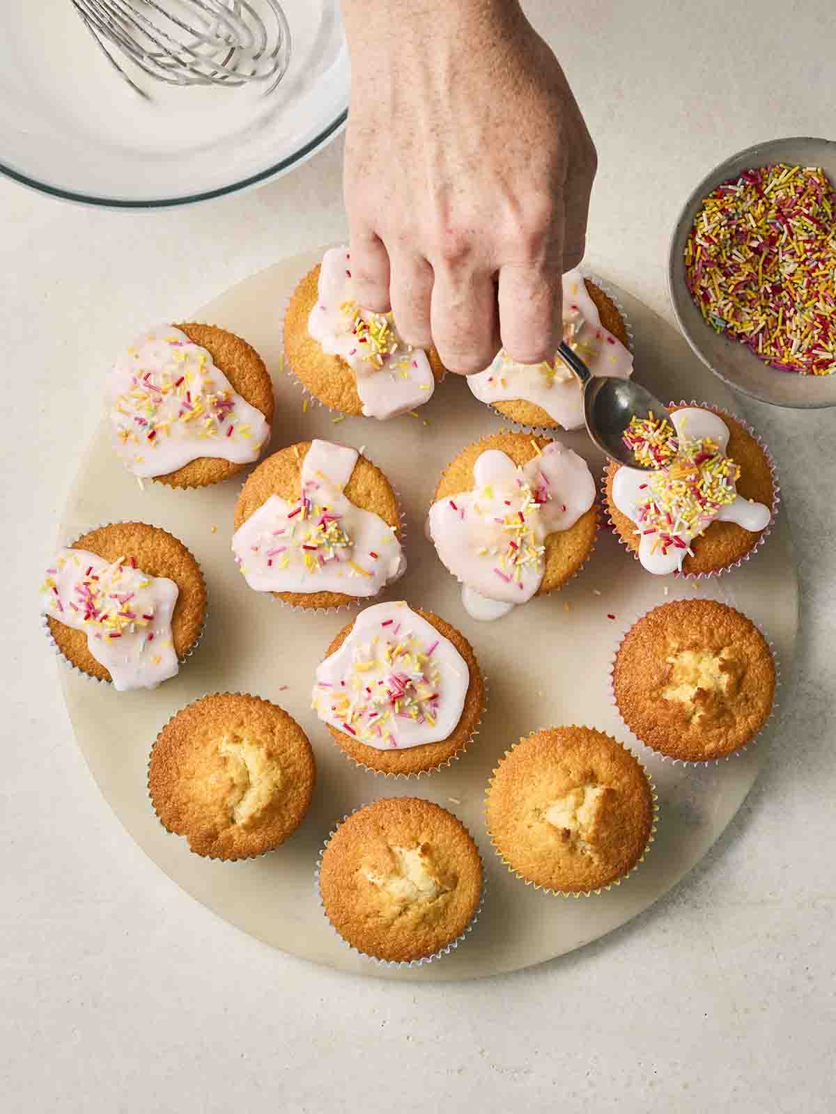 A hand sprinkling hundreds and thousands over the top of a plate of iced fairy cakes.