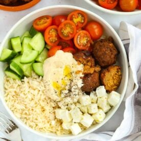 A white bowl filled with a Hummus Bowl salad, with rice, falafel, hummus, tomatoes and cucumber.