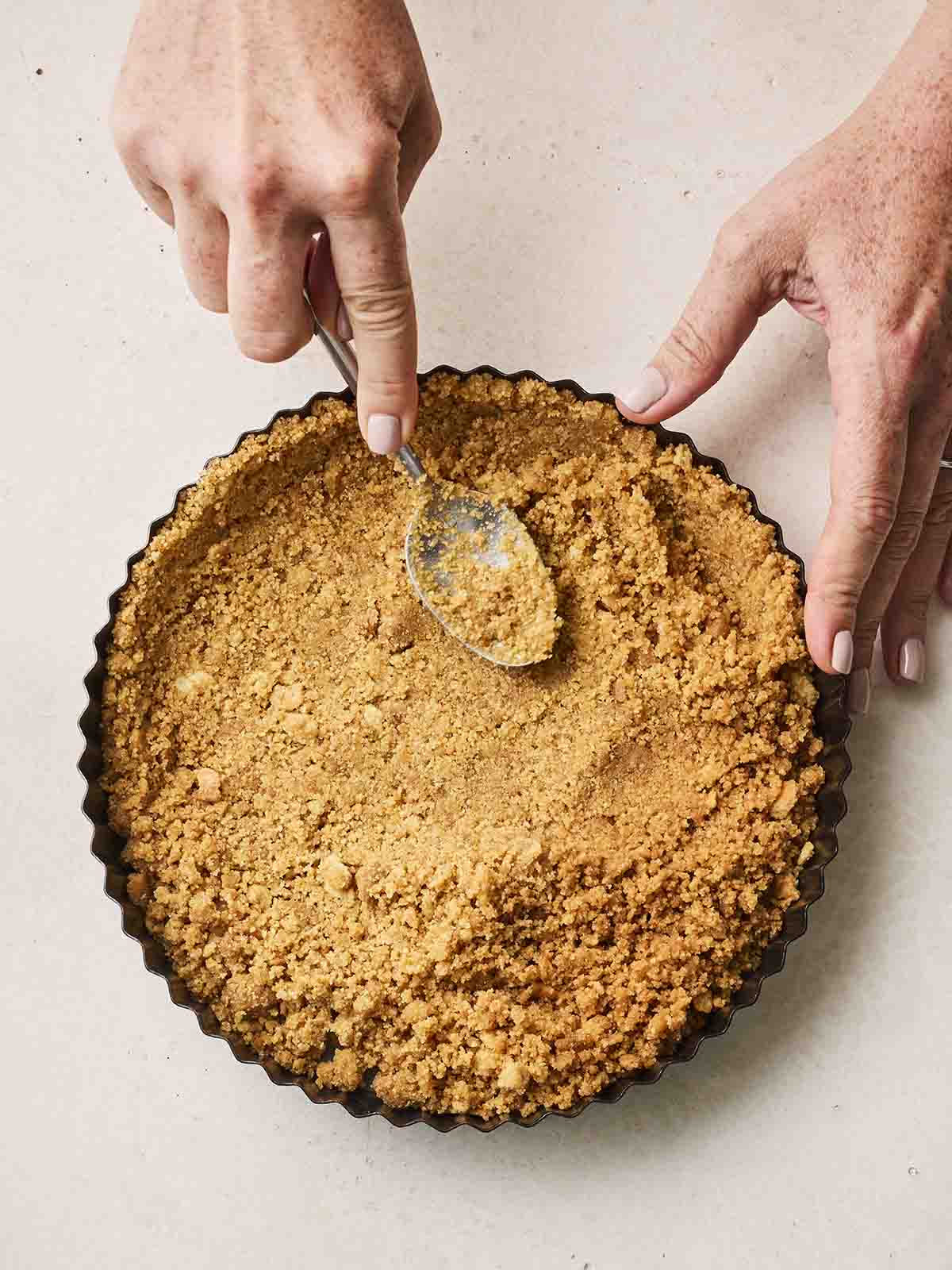 Crushed digestive biscuits being flatted into a flan tin with two hands holding a spoon.