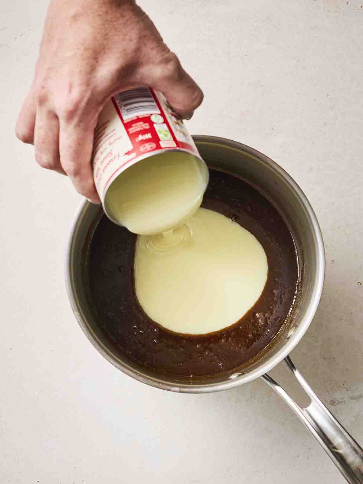 Condensed milk being added to a pan of brown liquid for making caramel, with a hand pouring it in.