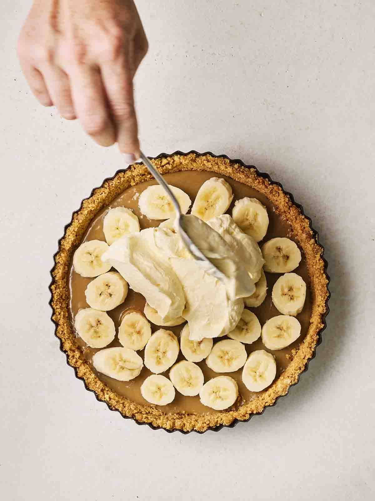 A dollop of whipped cream being put on top of a half-assembled Banoffee Pie with a hand spooning it on.