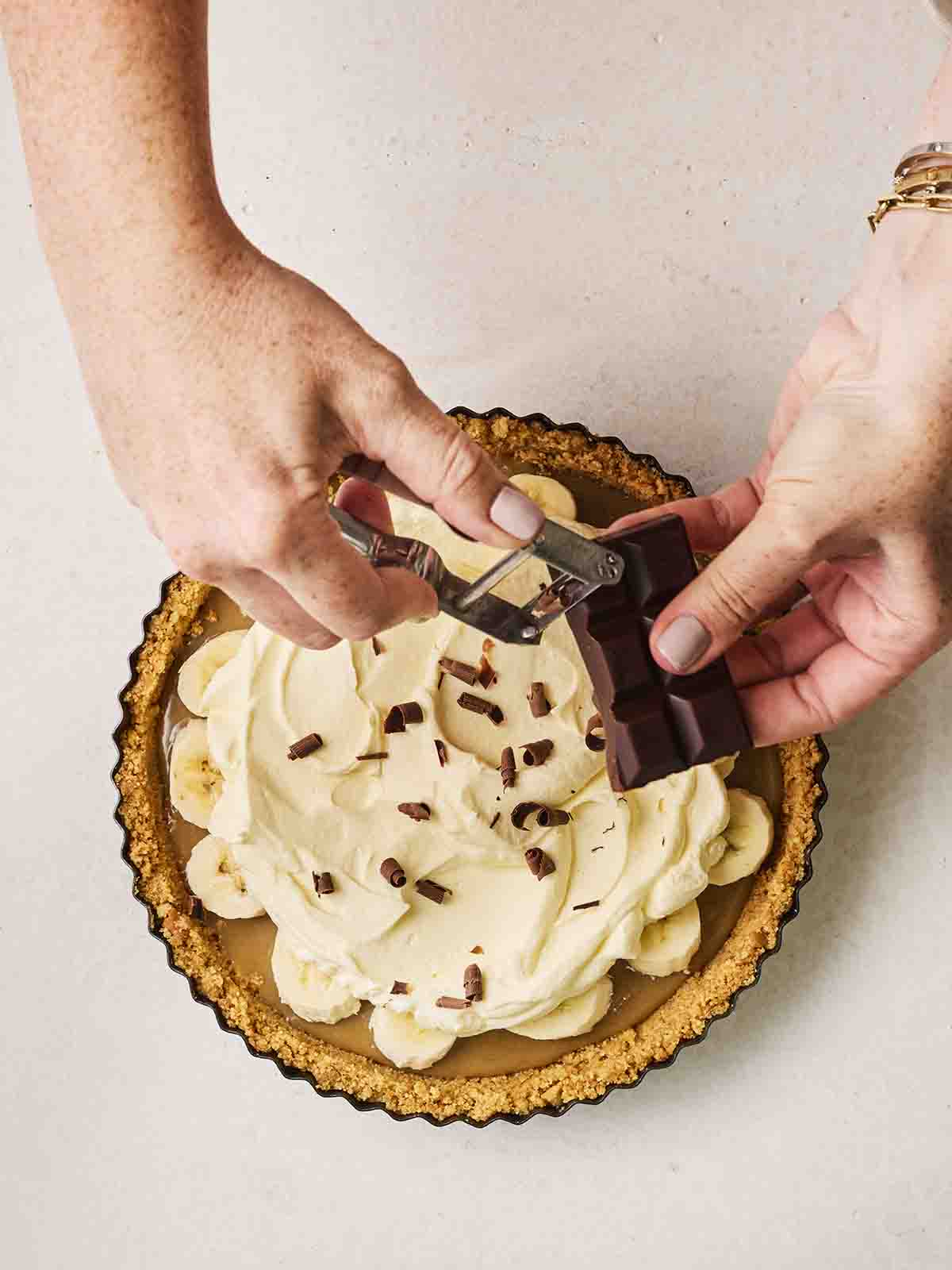 Two hands grating chocolate over a finished Banoffee Pie.