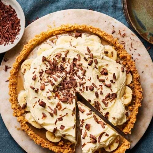 A homemade Banoffee Pie on a plate from above, with a slice cut out, on a blue tablecloth.