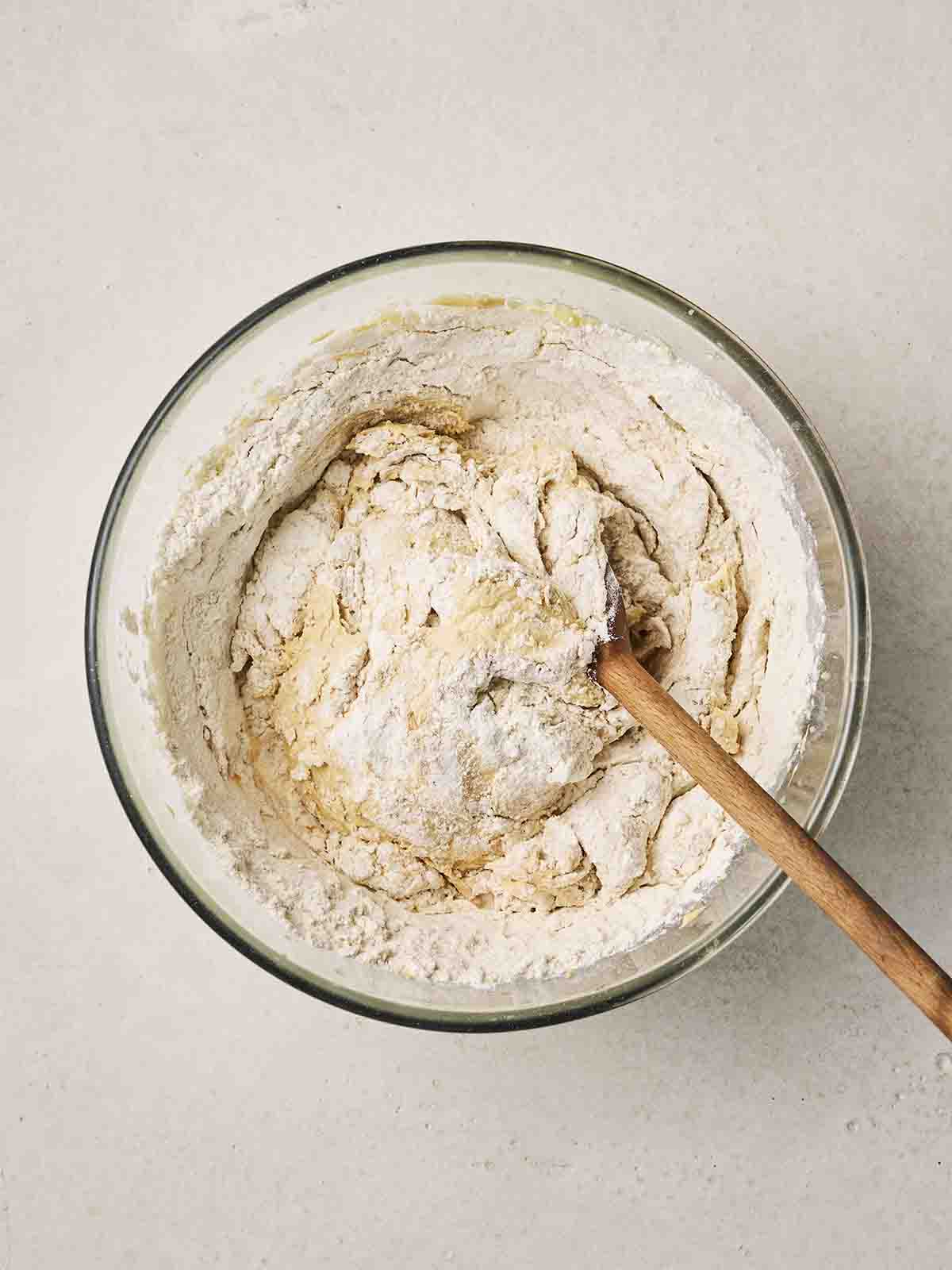Flour being mixed into cake batter in a glass bowl with a wooden spoon.