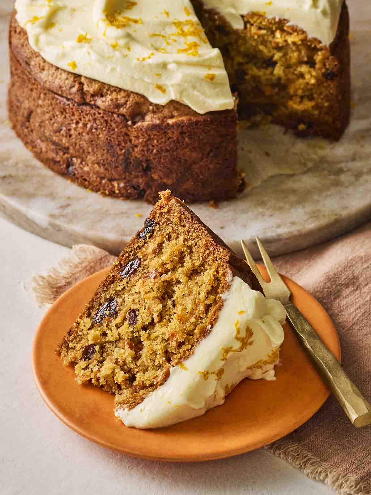 A slice of carrot cake on an orange plate with a cake fork, ready otherwise eat, with the rest of the cake in the background.