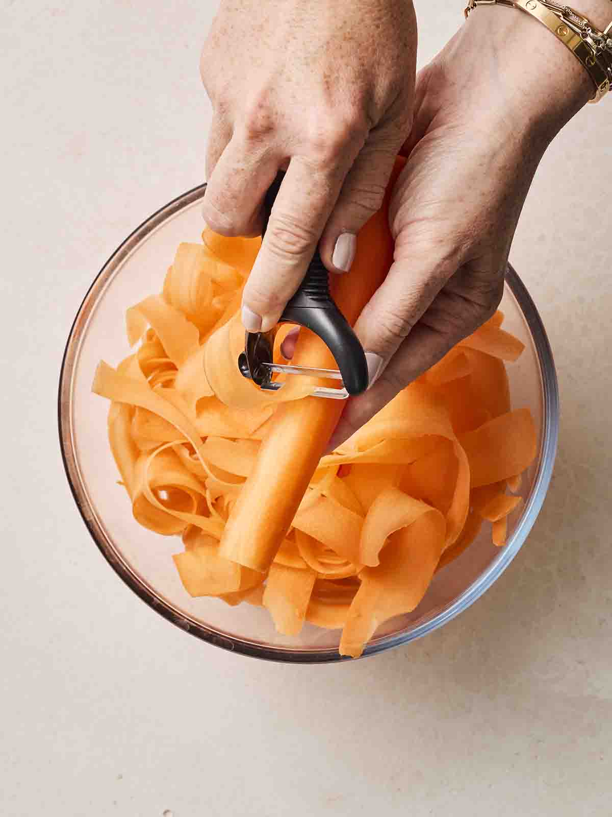 Two hands over a glass bowl, peeling carrots into ribbons for a Carrot Salad.