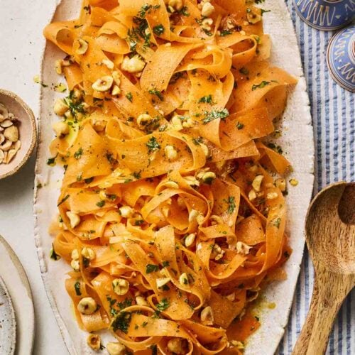 A platter of carrot salad with hazelnuts on a table with wooden utensils, salt and pepper and a blue and white cloth.