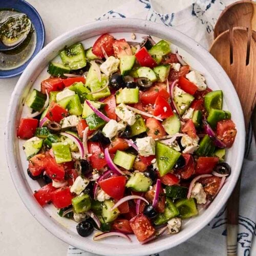 A plate of salad with peppers, tomatoes, cucumber, onion and feta for a Greek-style dish on a table with wooden utensils and dressing.