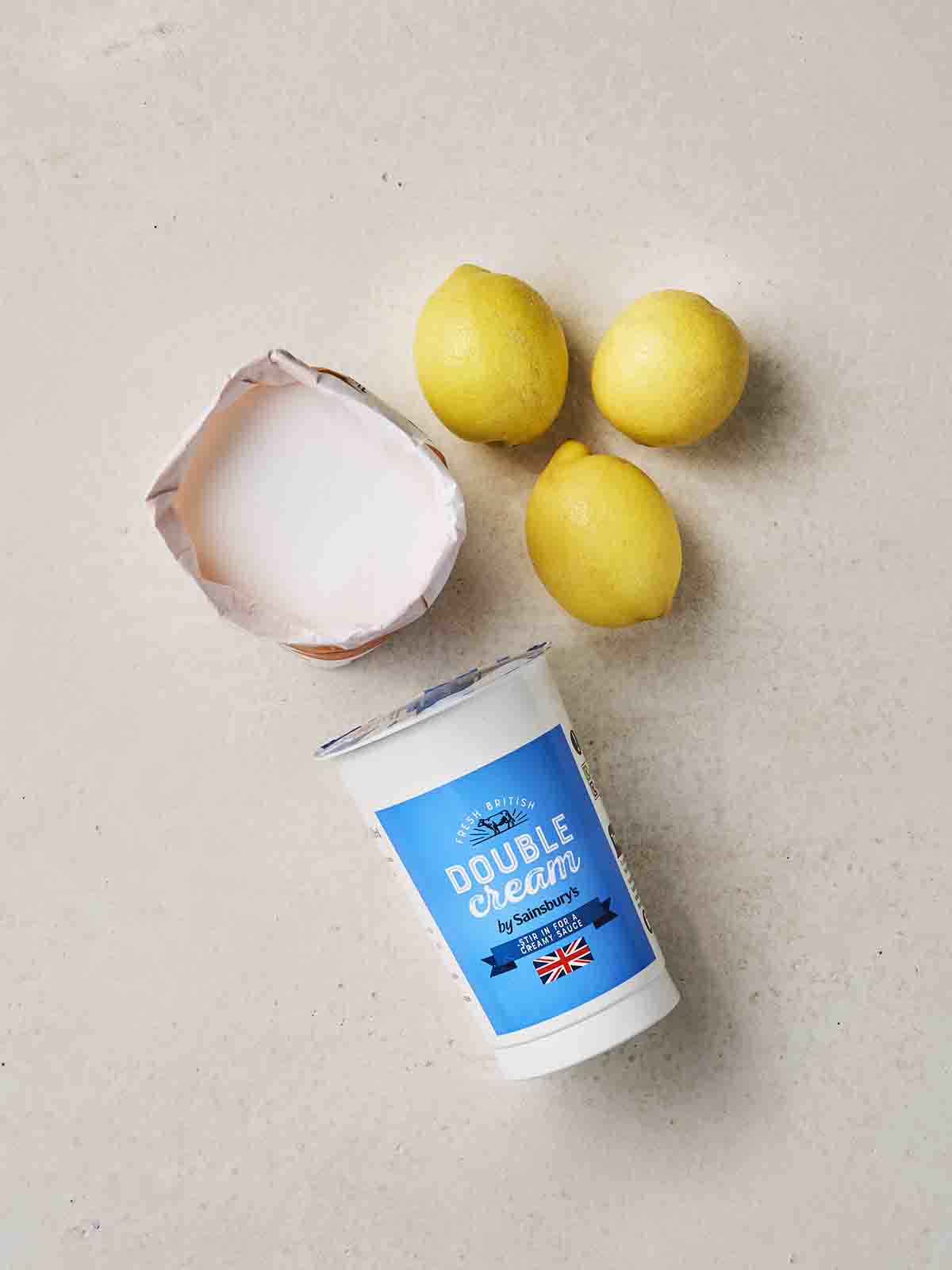 The three ingredients that make up a Lemon Posset recipe laid out on a white counter - lemons, sugar and cream.