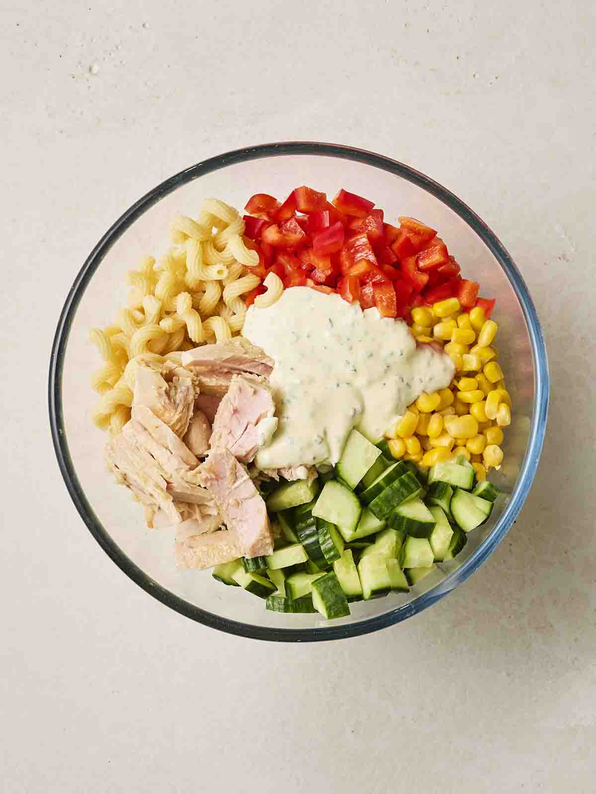 A glass bowl with Tuna Pasta Salad ingredients together, ready to mix, with dressing on top.