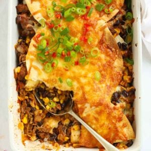 Looking down on a oven dish filled with Mexican beef with sweetcorn and beans, topped with tortillas and cheese, with a spoon dishing out.
