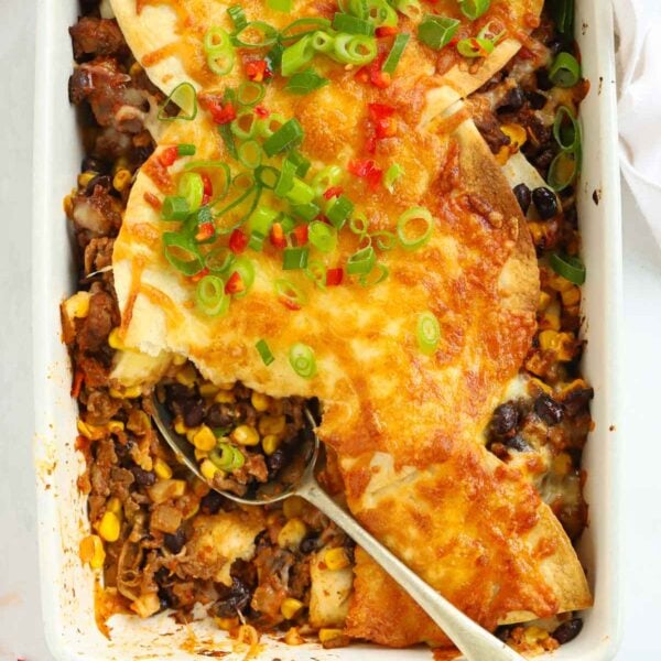 Looking down on a oven dish filled with Mexican beef with sweetcorn and beans, topped with tortillas and cheese, with a spoon dishing out.