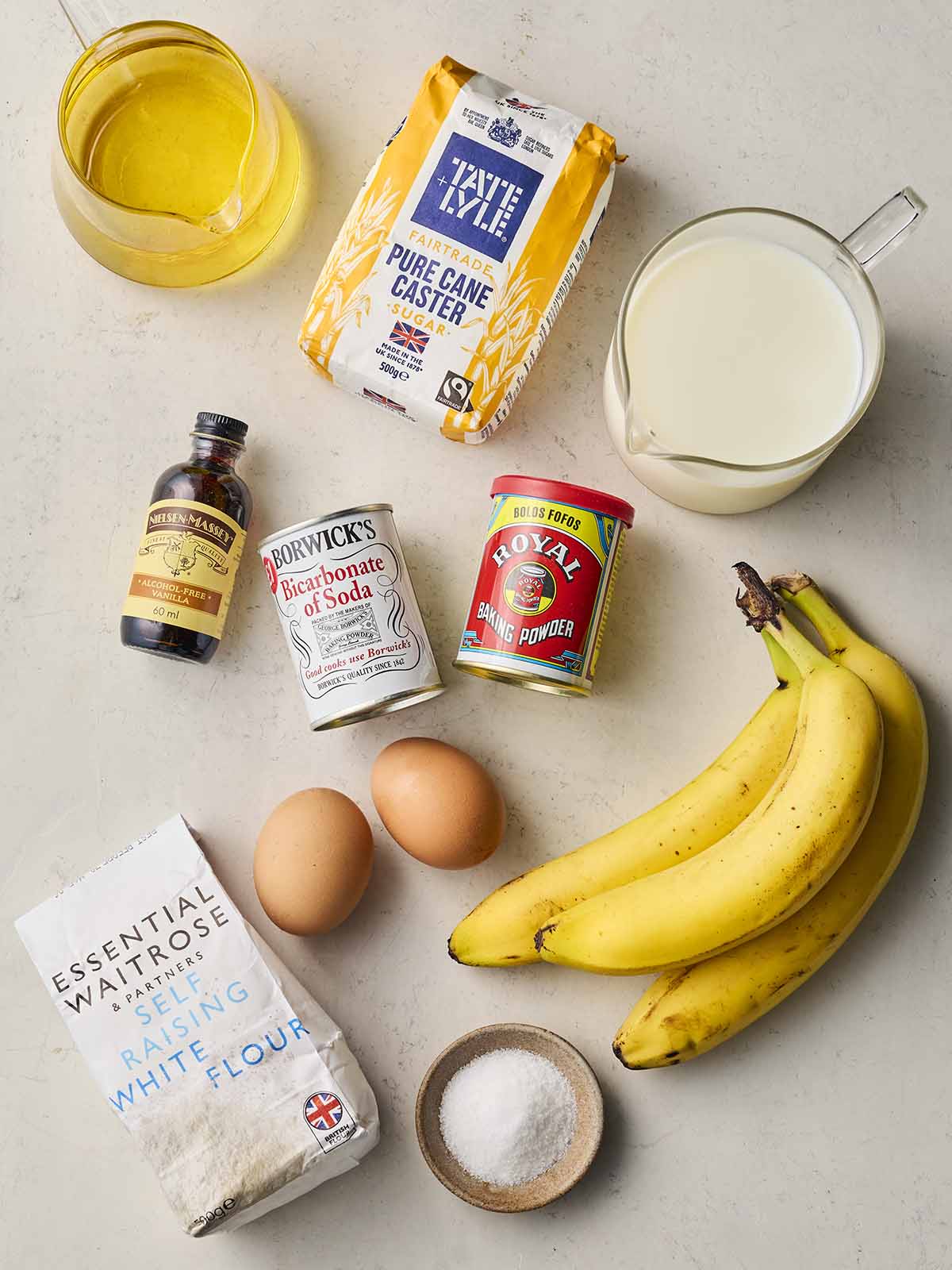 An assortment of ingredients on a white counter top, including bananas, milk, sugar, eggs, flour and oil.
