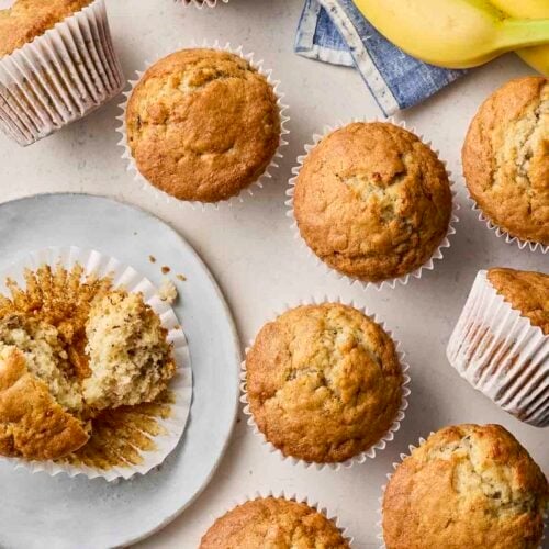 Lots of muffins on a table, with one half eaten on a plate and bananas to the side.