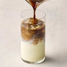 A glass of milk with ice, with coffee being poured into it.