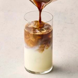 A glass of milk with ice, with coffee being poured into it.