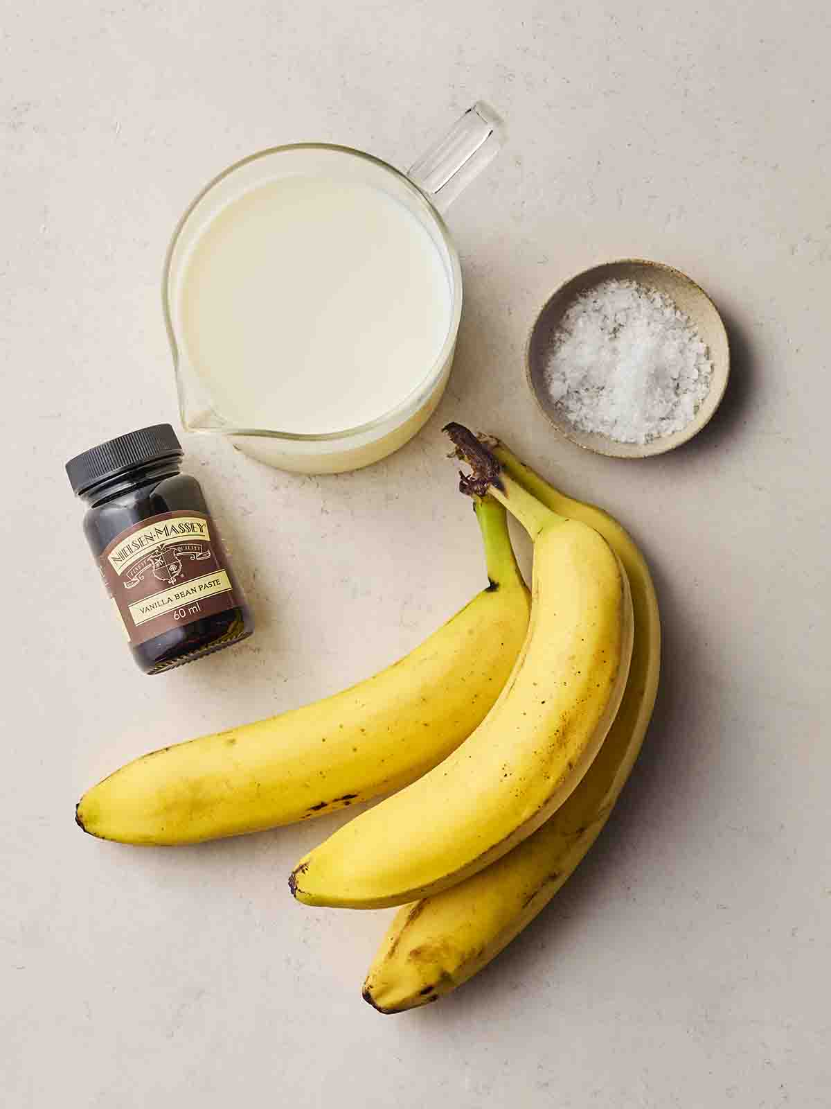 Ingredients laid out on a white counter, including bananas, vanilla, salt and milk.