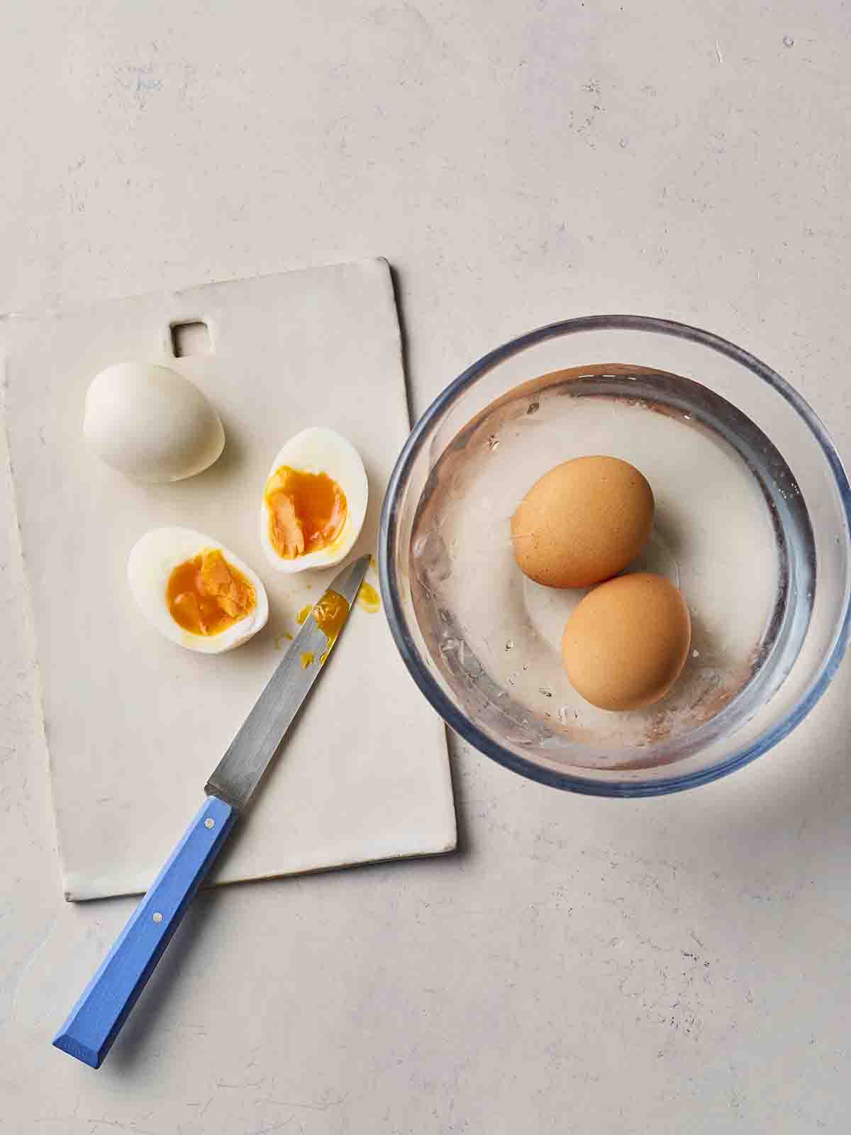 Two boiled eggs in a glass bowl with water, and two eggs on a chopping board, one cut in half, with a knife next to it.