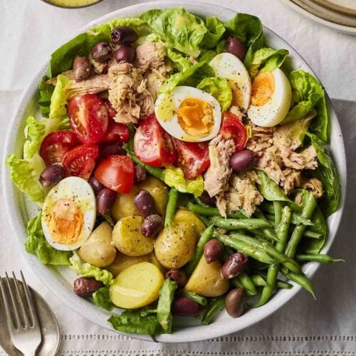 A big plate of salad on a white table cloth, with dressing and cutlery to the side.