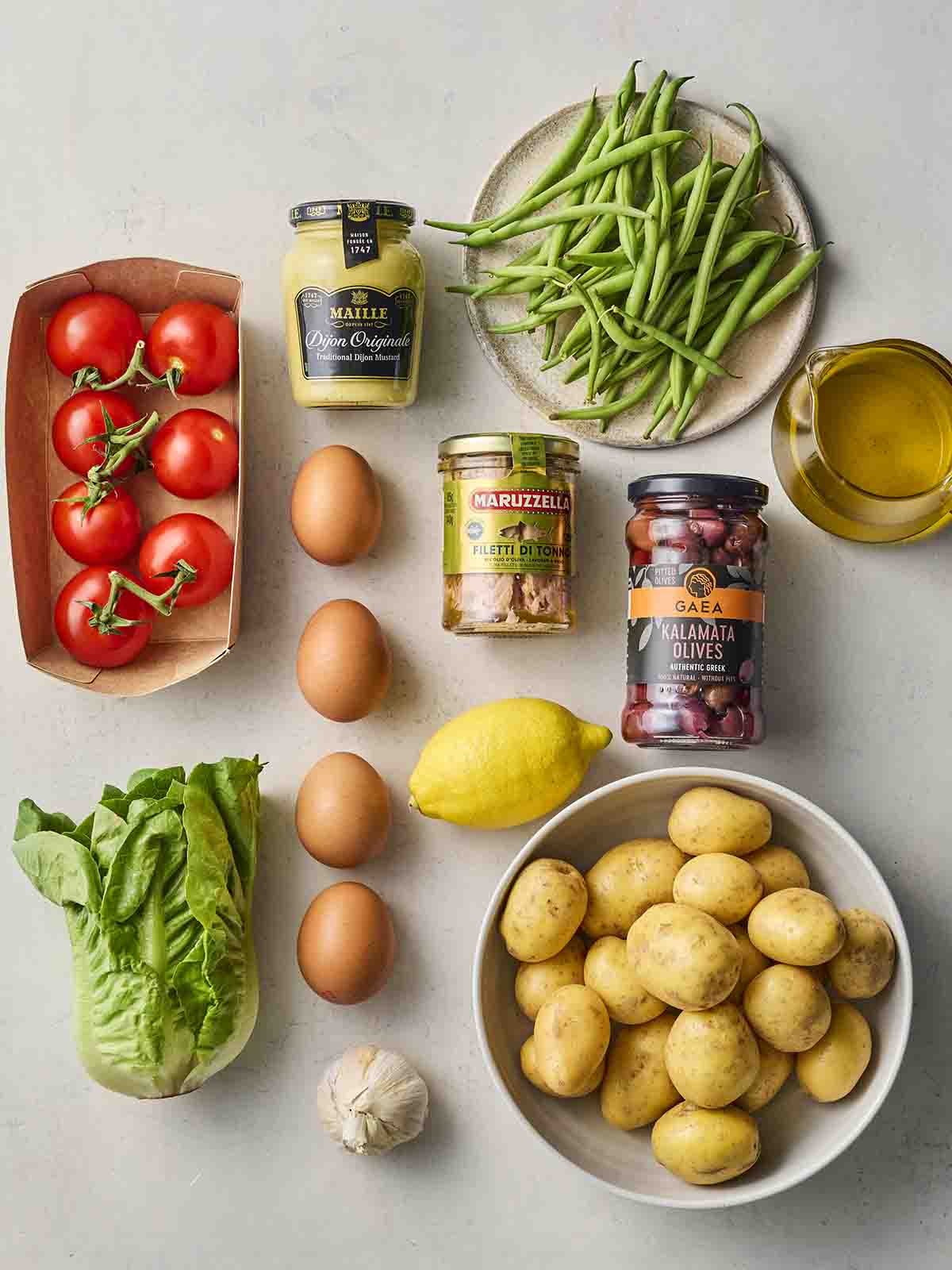 Ingredients laid out on a white counter, including potatoes, eggs, tuna, olives, green beans, lettuce and tomatoes.